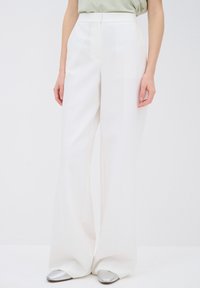 Taille fine, pantalon blanc à jambes larges porté avec un haut vert pâle et des chaussures argentées pointues, visible de la taille jusqu’aux pieds.