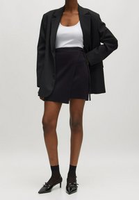 Blazer nero su una canotta bianca, abbinato a una gonna avvolgente scura. La modella indossa tacchi a spillo neri e calze nere. Texture lisce.