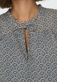 Blouse à motifs en bleu clair avec des designs géométriques en brun et bleu foncé. Présente une fermeture à nouer et un col froncé. Tissu texturé.