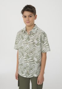 Camicia a maniche corte di colore verde chiaro con motivi di foglie bianche, con bottoni sul davanti e colletto, realizzata in tessuto leggero. Abbinata a pantaloni verde oliva.
