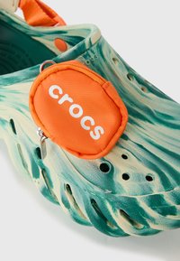 Crocs à motifs verts et beiges avec un design arrondi, des trous de ventilation et une pochette orange avec un logo blanc attaché.