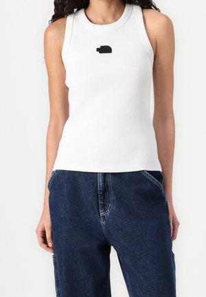 Frau trägt ärmelloses weißes Rippentop mit kleinem schwarzem Logo auf der Brust und lockere dunkelblaue Jeans mit Händen in den Taschen.
