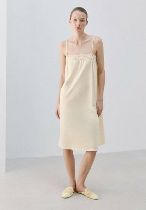 Kvinde i en lysegul ærmeløs kjole med blondekant og matchende slip-on vævede sko, stående mod en ensfarvet lys baggrund.