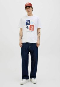 Hvid T-shirt med et farverigt grafisk design af blade i blå, rød og creme. Bæres med løstsiddende mørke denim jeans og hvide sneakers.