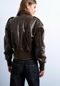 Femme portant une veste en cuir marron foncé coupée, avec des poignets et un ourlet côtelés, associée à un jean en denim foncé taille haute, de dos.