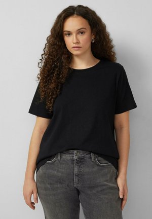 Frau mit langen lockigen Haaren, die ein schlichtes schwarzes T-Shirt mit kurzen Ärmeln und graue Jeans vor einem grauen Hintergrund trägt.