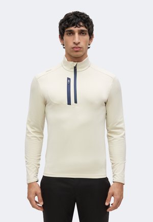 Junger Mann mit dunklem lockigem Haar, der ein hellbeiges Langarmshirt mit Reißverschlusstasche auf der Brust und schwarze Hosen trägt und aufrecht steht.