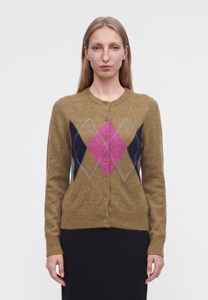 Femme aux cheveux blonds raides porte un cardigan marron boutonné avec un motif losanges rose et bleu foncé, associé à une jupe noire.