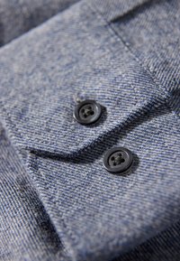 Jeansskjortstyg i ljusblått med svaga ljusare ränder, med två svarta knappar och en spetsig krage. Texturerad ytdetalj synlig.