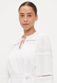 Vestido branco com detalhes em renda, gola rodada e mangas longas. Apresenta um fecho atado ao pescoço e um cinto atado à cintura.