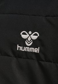 Schwarzer gepolsterter Stoff mit horizontalen Nähten zeigt ein weiß besticktes Logo von "hummel" mit einem charakteristischen Symbol über dem Text.