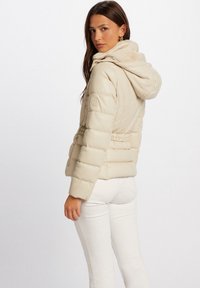 Giacca puffer beige con vita regolabile e cappuccio. Presenta un design trapuntato e una patch con logo sul maniche. Indossata con pantaloni bianchi.