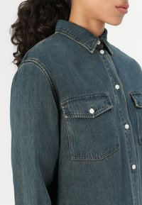 Denimhemd in Mittelblau mit Knopfleiste, spitzem Kragen und zwei Brusttaschen. Verfügt über weiße Knöpfe und eine lockere Passform.