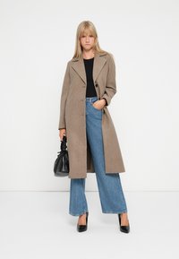Long manteau beige avec un col cranté, fermeture à un bouton, porté sur un haut noir et un jean bleu à jambes larges, assorti à un sac à main noir.