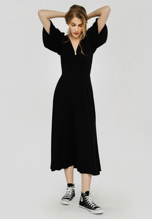 Femme en robe midi noire à manches courtes larges et baskets montantes noires, debout avec les mains derrière la tête sur fond gris.