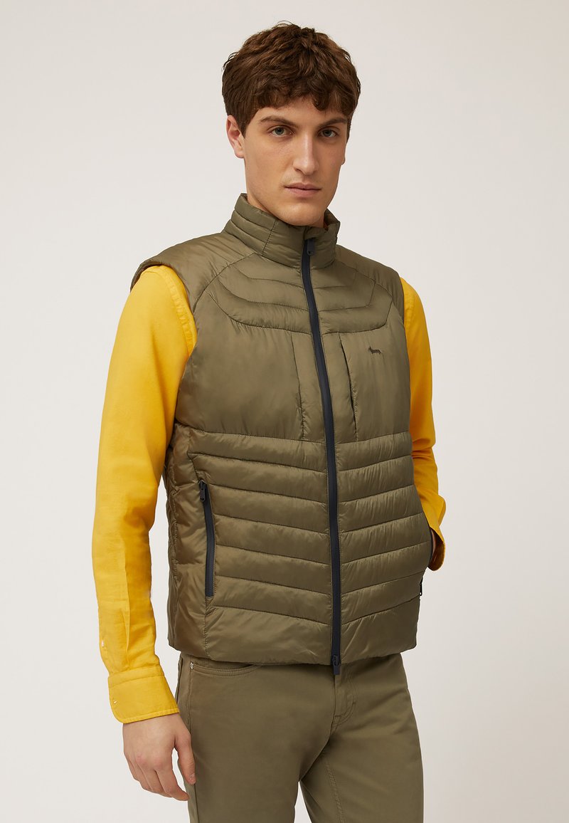 Chaleco acolchado verde oliva con cuello alto, cierre frontal con cremallera y bolsillos laterales. Lleva una camisa de manga larga amarilla debajo. Textura suave.