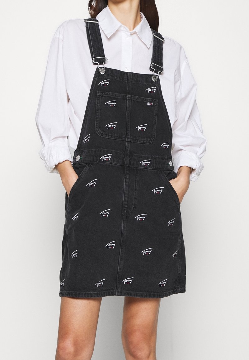 Kvinna som bär svart jeansoverallklänning med vita broderade logotyper, kombinerad med vit långärmad skjorta med knappar, händerna i fickorna.