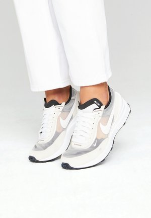 Osoba nosząca białe i szare trampki Nike z czarnymi podeszwami i białymi sznurowadłami, w połączeniu z białymi krótkimi spodniami na jednolitym tle.
