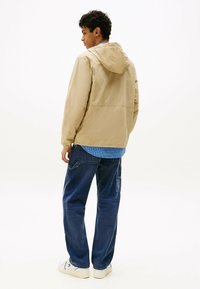 Jasno beżowa kurtka z kapturem i elastycznymi mankietami, noszona na niebieskiej koszuli w kratę oraz ciemnoniebieskich luźnych dżinsach. Całość dopełniają białe trampki.