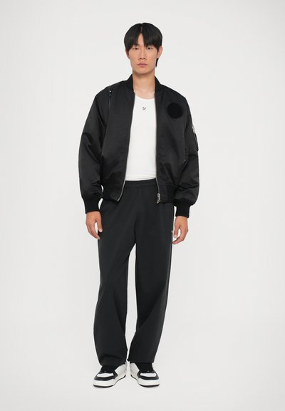Sort bomberjakke med lynlåslukning og sidelommer, båret over en hvid tætsiddende skjorte og sorte sweatpants. Sorte og hvide sneakers.