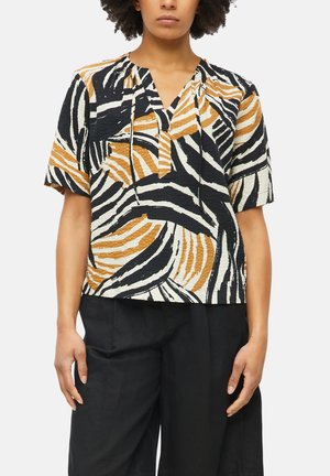Femme portant un chemisier à manches courtes avec des rayures abstraites noires, blanches et moutarde et un pantalon noir taille haute sur fond blanc.