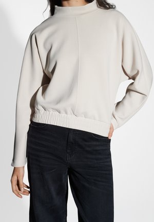 Femme portant un haut beige clair à manches longues avec une taille élastique et un jean noir taille haute, debout avec une main sur la hanche.
