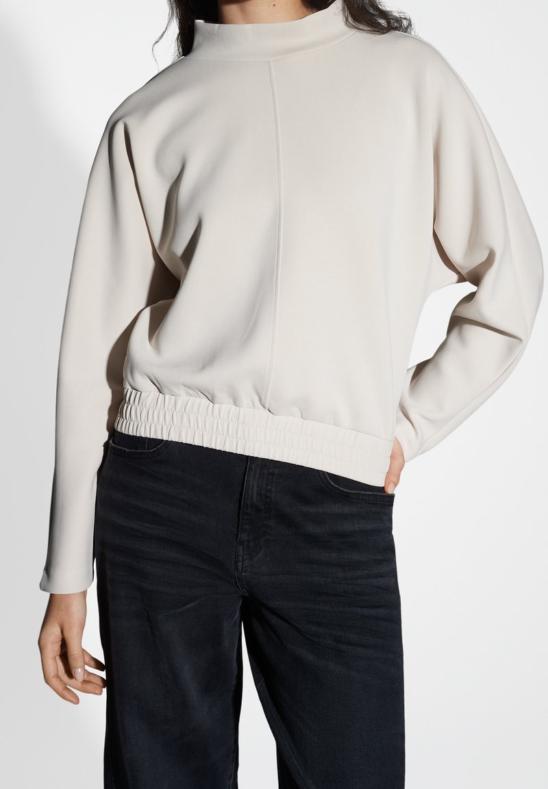 Femme portant un haut beige clair à manches longues avec une taille élastique et un jean noir taille haute, debout avec une main sur la hanche.