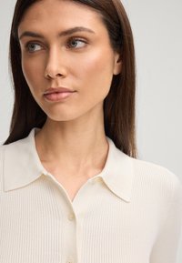 Ragazza giovane con capelli castani lisci, che indossa una camicia a coste button-up color avorio, mentre guarda a sinistra contro uno sfondo semplice.