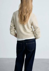 Beigen sävyinen neulottu neule rentolla istuvuudella ja käärittyinä hihoina, yhdistetty tummansinisiin korkeavyötäröisiin farkkuihin, joissa on takataskut.