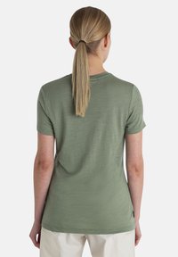 T-shirt verde oliva a maniche corte con scollatura rotonda e una texture morbida, presenta un orlo curvo e una vestibilità ampia, indossato con pantaloni chiari.