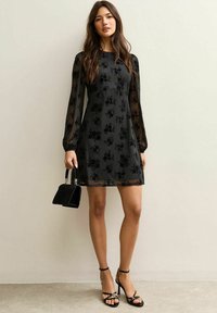 Czarna sukienka w kwiatowy wzór, przezroczyste długie rękawy i dopasowana góra. Zestawiona z czarnymi sandałami na obcasie i małą czarną torebką.