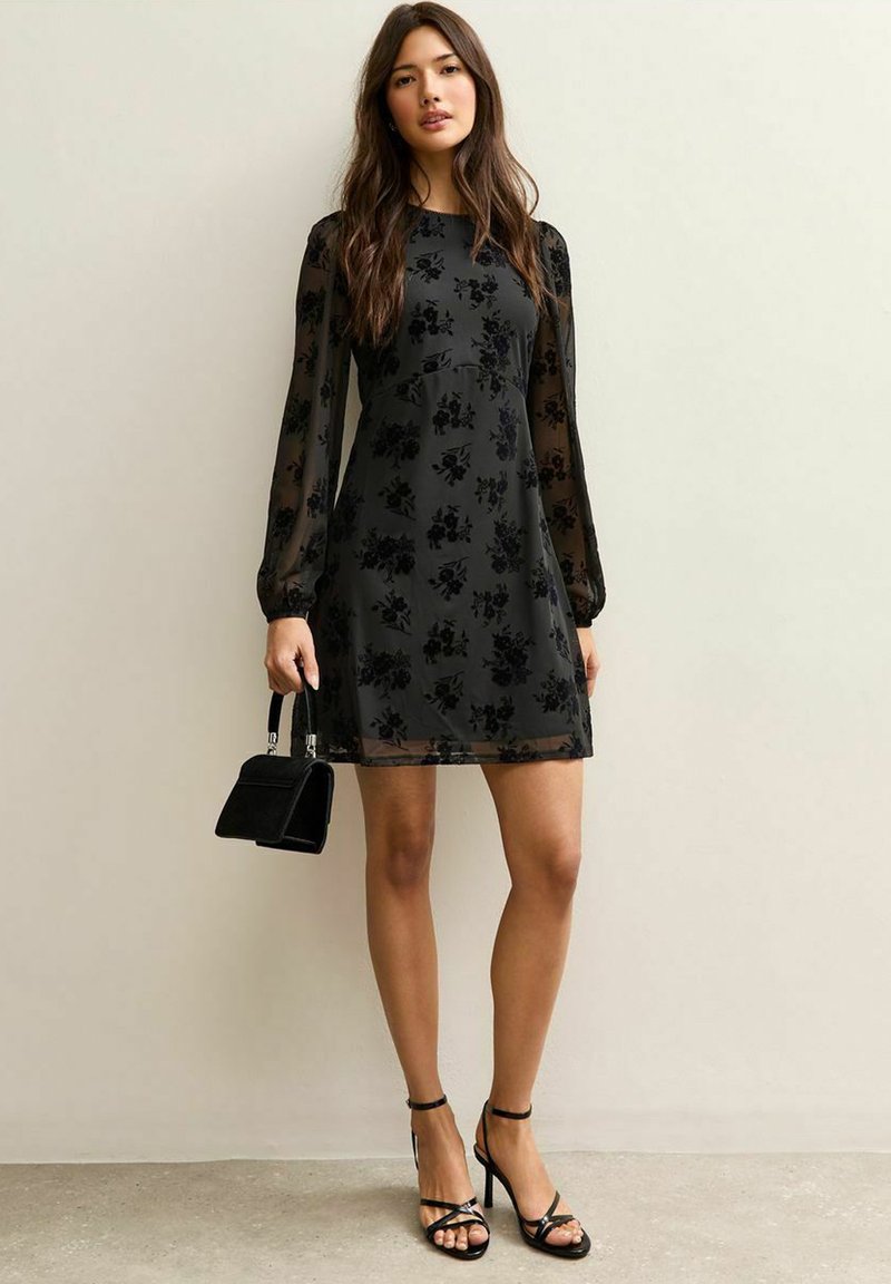 Czarna sukienka w kwiatowy wzór, przezroczyste długie rękawy i dopasowana góra. Zestawiona z czarnymi sandałami na obcasie i małą czarną torebką.