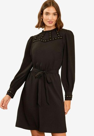 Abito nero con maniche lunghe, scollatura arricciata e vita aderente. Presenta borchie decorative dorate sulle spalle e sui polsini.