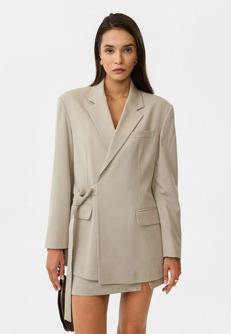 Beige op maat gemaakte blazer met een knoopdetail in de taille, een enkele borstzak en een soepele textuur, gecombineerd met een bijpassende minirok.