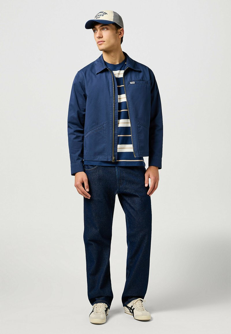 Granatowa kurtka z zamkiem z przodu i kieszeniami po bokach, nałożona na pasiasty granatowo-biały t-shirt. Ciemne dżinsy i kremowe sneakersy dopełniają całość.