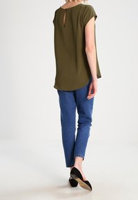 Blouse vert olive à manches courtes, décolleté rond et détail en forme de trou dans le dos. Associée à un jean bleu et des chaussures noires. Texture lisse, coupe ample.