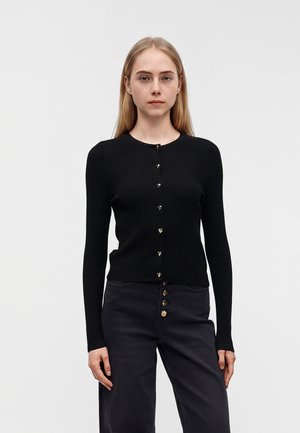 Femme portant un cardigan noir côtelé boutonné et un pantalon sombre taille haute, debout devant un fond clair uni.