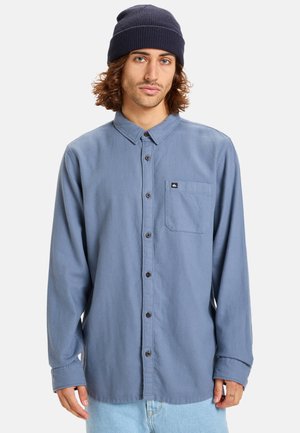 Langärmliges Button-Up-Hemd aus hellblauem Stoff, ausgestattet mit einer Brusttasche und braunen Knöpfen. Tragen mit einer marineblauen Strickmütze.