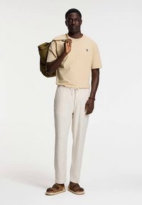 Bež majica s kratkim rukavima, svijetle hlače na pruge i smeđe sandale. Model drži torbu maslinasto zelene boje. Ležerna, opuštena kroj s vezicom u struku.