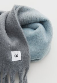 Sciarpa di lana morbida con una sfumatura dal blu chiaro al grigio, con dettagli in frangia e un'etichetta bianca con logo. Texture complessiva liscia.