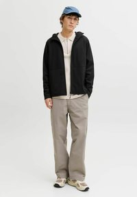 Veste noire zippée portée sur un polo beige, associée à un pantalon beige clair et des baskets crème. Casquette bleue et détails subtils de texture visibles.