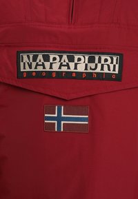 Rød stof med et fremtrædende sort og hvidt "NAPAPIJRI geographic" mærke. Har et broderet norsk flag med brun baggrund og blå detaljer.