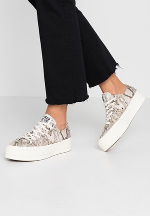 Stopy w beżowych sneakersach na platformie z motywem węża, białą podeszwą i sznurówkami, zestawione z czarnymi skróconymi dżinsami z postrzępionymi brzegami.