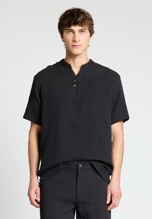 Young man with short brown hair wearing a loose black short-sleeve shirt with three buttons and matching black pants, standing against a plain background.