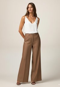 Pantalons amples marron avec un motif texturé, assortis à un haut sans manches blanc avec un décolleté en V. Sandales noires à brides.