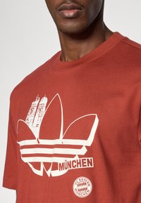 Μπλουζάκι από βαμβάκι χρώματος σκουριάς με λευκό λογότυπο Adidas με τρεις ρίγες, ορίζοντα του Μονάχου κα ι έμβλημα της FC Bayern München.