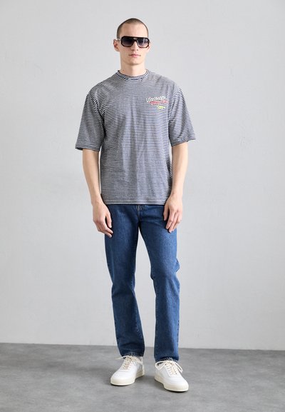 Tricou cu mânecă scurtă, în dungi alb-negru, asociat cu blugi negri și adidași albi. Detalii grafice îndrăznețe pe tricou.