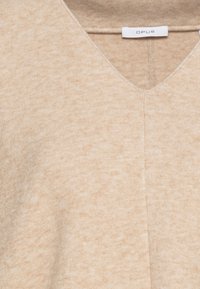 Pull beige en tricot avec un col en V, présentant une texture douce et un motif subtil. L'étiquette indique "OPUS", avec un design épuré et minimaliste.