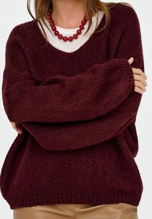 Woman wearing oversized dark burgundy knit sweater, white shirt underneath, red bead necklace, and beige pants with arms crossed.