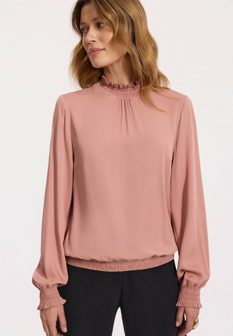 Różowa bluzka z długim rękawem, z falbaniastym wysokim kołnierzem, marszczonymi mankietami i luźnym krojem. Wykonana z gładkiego, lekkiego materiału.